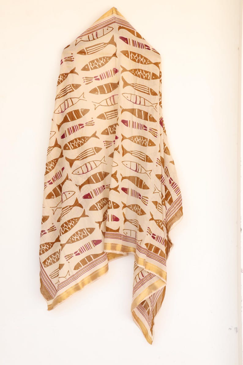 May include: A beige scarf with a repeating pattern of brown fish illustrations. The scarf has a gold border with a thin maroon stripe. The fabric is draped, showcasing the design and texture.