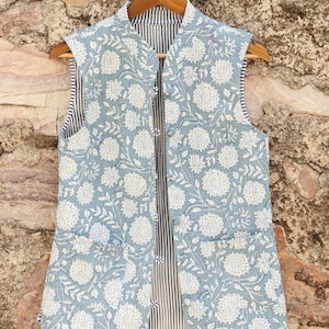 May include: A light blue floral print vest with a white floral pattern. The vest has a striped lining and two front pockets. It is hanging on a wooden hanger against a stone wall.