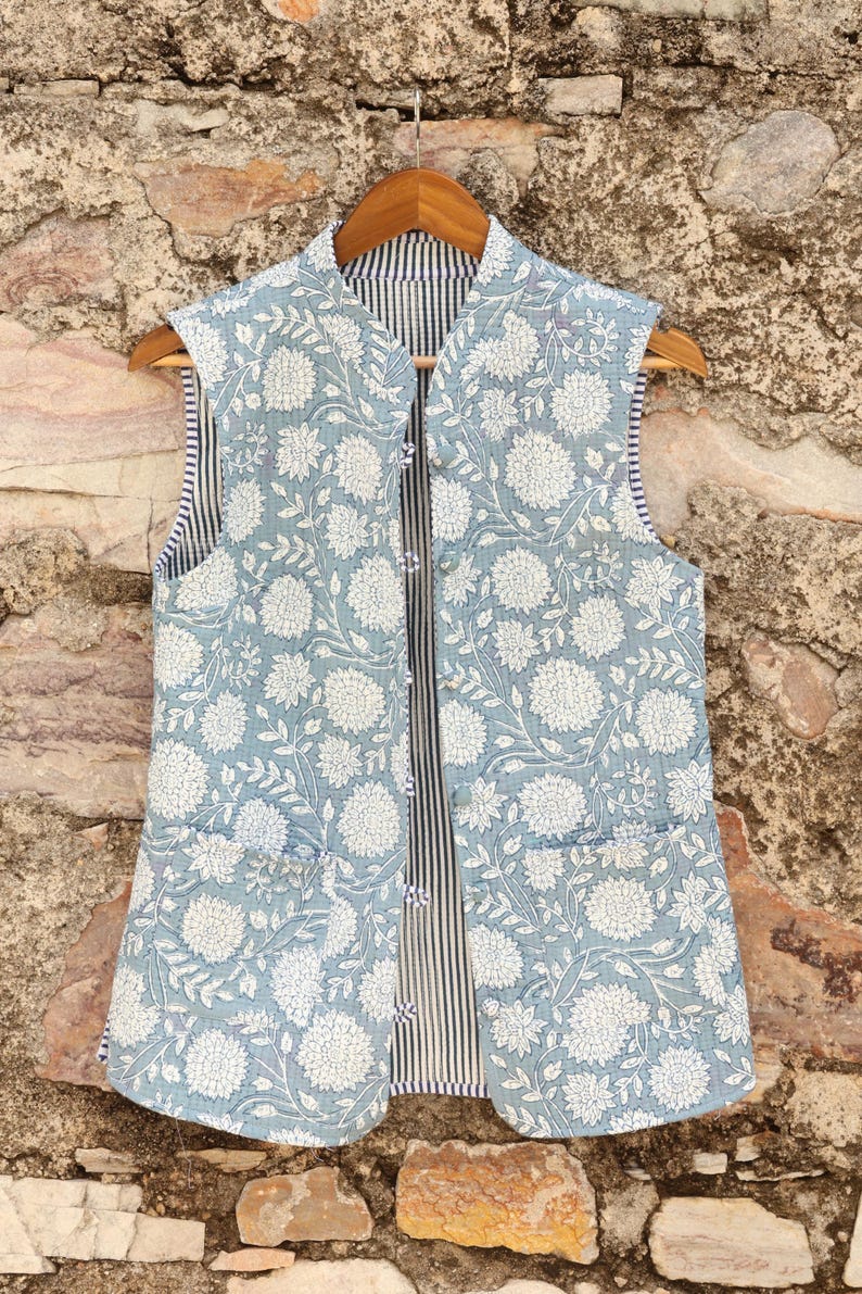 May include: A light blue floral print vest with a white floral pattern. The vest has a striped lining and two front pockets. It is hanging on a wooden hanger against a stone wall.