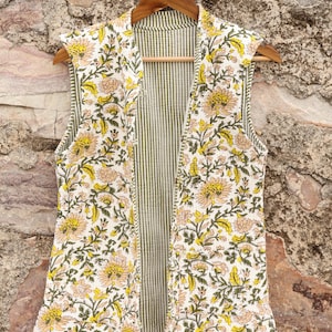 May include: A sleeveless, open-front vest with a floral pattern in yellow and peach on a white background. The interior lining features vertical green and white stripes. The vest is hanging on a wooden hanger.