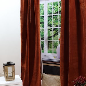 May include: Two rust-colored velvet curtains hang from a silver curtain rod in front of a window with a view of green trees. A white nightstand with two drawers is in the foreground, along with a decorative lantern and a white throw pillow.