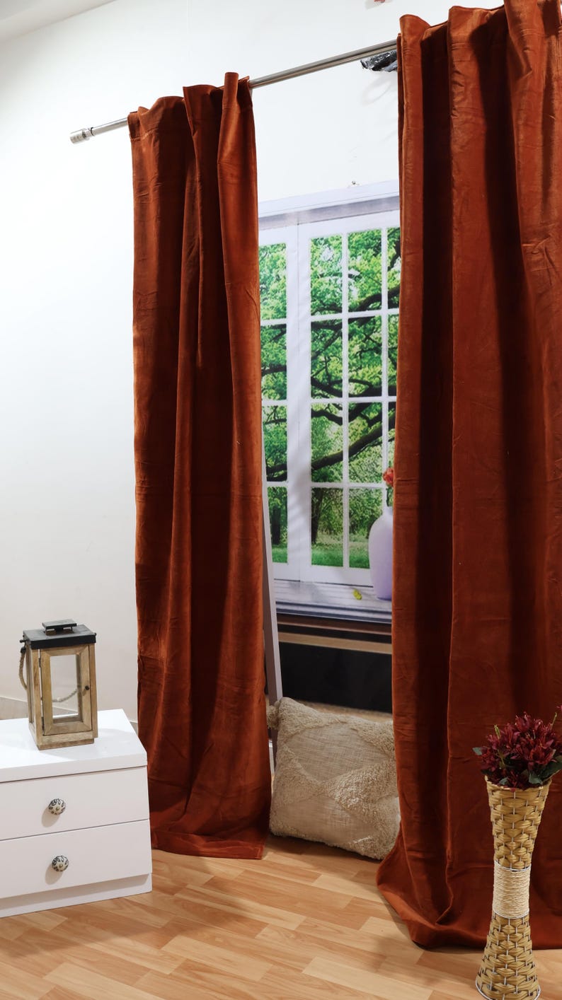 May include: Two rust-colored velvet curtains hang from a silver curtain rod in front of a window with a view of green trees. A white nightstand with two drawers is in the foreground, along with a decorative lantern and a white throw pillow.