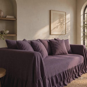 May include: A deep purple sofa cover featuring a double-layered ruffled skirt along the base. The cover is made of a textured fabric and includes matching square throw pillows arranged across the back, providing a full drape over the sofa frame.