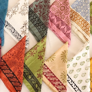 Puede incluir: Una colección de pañuelos estampados de colores. Cada pañuelo está doblado en forma de triángulo, mostrando varios diseños florales, paisley y geométricos. La paleta de colores incluye tonos de azul, naranja, verde, rosa y amarillo.