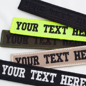 May include: Five different color fabric patches with black stitching. The patches are rectangular and have the text "YOUR TEXT HERE" embroidered on them. The colors of the patches are black, neon green, olive green, tan, and black with white text.