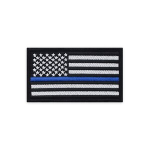 以下が含まれることがあります： 中央に細い青い線が引かれたアメリカ国旗の刺繍パッチ。旗は黒地に白と青です。