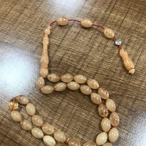 May include: A beige prayer beads necklace with a silver charm. The beads are oval shaped and have a translucent appearance. The necklace is strung with a red cord.