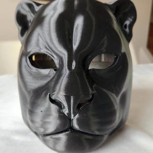 May include: A black panther mask with eye openings and a detailed nose and mouth. The mask is made of a textured material and is designed to fit the shape of a human face. The mask is a solid black color.