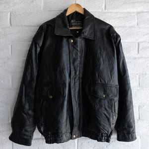 Könnte beinhalten: Schwarze Lederjacke mit klassischem Design, mit Kragen, zwei Vordertaschen und Knopfverschluss. Die Jacke hängt an einem Holzbügel vor einer weißen Backsteinmauer. Das Etikett lautet "GENUINE LEATHER".