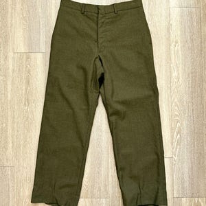 Puede incluir: Un pantalón de color verde oliva con una cintura estructurada con trabillas para cinturón, una cremallera central y un cierre de botón. El pantalón tiene un corte de pierna recta con un acabado sólido y una costura visible en el centro.
