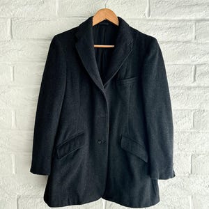 Puede incluir: Un abrigo de lana gris oscuro con un diseño clásico, colgado de una percha de madera. El abrigo presenta una solapa con muescas, dos bolsillos delanteros y un cierre de un solo botón. El abrigo se exhibe contra una pared de ladrillos blancos.