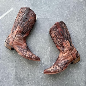 Op de afbeelding: Een paar bruine leren cowboylaarzen met ingewikkelde donkerbruine gestikte overlays en borduursels op de schacht en de neus. De laarzen hebben een puntige neus, een gestapelde houten hak en treklussen aan de bovenkant van de schacht.