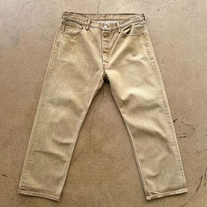 May include: Tan-colored denim jeans with a button fly and classic five-pocket design. The jeans are laid flat on a concrete surface, showcasing their straight-leg cut and faded appearance. A casual, everyday wear item.