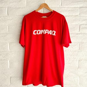 Peut inclure: Un t-shirt rouge avec le mot "COMPAQ" en blanc imprimé sur le devant. Le t-shirt est suspendu à un cintre en bois contre un mur de briques blanches.