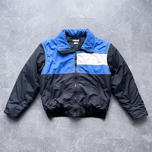 May include: A color-blocked puffer jacket featuring a black lower section and sleeves, a blue upper chest and collar, and a white horizontal stripe on the right chest. The jacket has a full-length front zipper and a black label reading ossi.