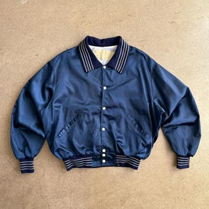 May include: A navy blue satin bomber jacket with white striped detailing on the collar, cuffs, and hem. The jacket has a button-up front and two front pockets. The cuffs and hem are ribbed.
