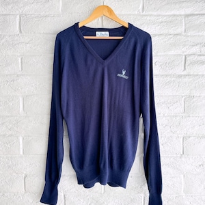 May include: Navy blue V-neck sweater with long sleeves. The sweater has a small embroidered design on the chest and a label at the neckline. The sweater is hanging on a wooden hanger against a white brick wall.