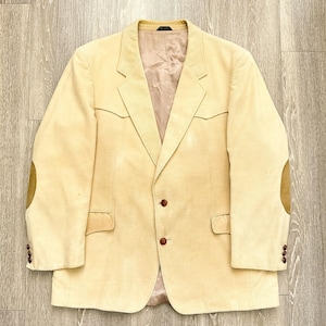 May include: A light yellow corduroy blazer with brown elbow patches and brown buttons. The blazer has a notched lapel and two front pockets. The interior lining is a light tan color. The blazer is laid flat on a wooden surface.