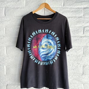 May include: Black t-shirt with a circular graphic featuring a red and blue design and the text "we can't dance" in yellow. The graphic is surrounded by small blue silhouettes of people. The shirt is hanging on a wooden hanger.