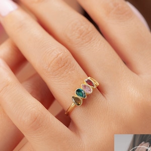 May include: Gold ring featuring a row of five pear-shaped gemstones in various colours. The gemstones are green, brown, pink, light pink, and red.