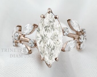 May include: A rose gold engagement ring with a large marquise-cut diamond center stone. The band features leaf and small diamond accents. The ring is set against a white background. The text "TRINETRA DIAMOND" is visible.