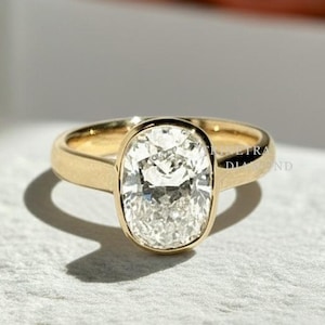 May include: A gold ring with a large, oval-shaped diamond. The diamond is set in a bezel setting, and the band is smooth and polished. The ring is displayed on a white surface.