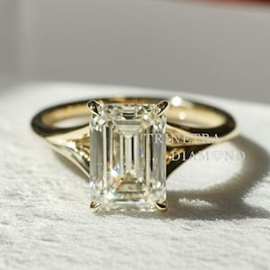 May include: A gold engagement ring with an emerald-cut diamond. The ring has a split shank design. The diamond is a light yellow color and is set in a four-prong setting. The ring is on a white surface.