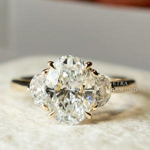 May include: A gold engagement ring with a large oval-cut diamond center stone and two smaller side stones. The ring is set on a white surface, with a blurred background. The ring is a classic and elegant design.