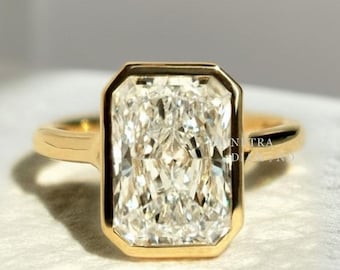 May include: A radiant-cut diamond ring with a gold band. The diamond is rectangular with a bezel setting. The ring is set on a white surface, with a blurred background of pink and white.