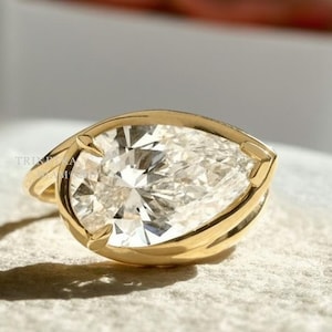 May include: A gold ring featuring a pear-shaped diamond. The diamond is set in a bezel setting, and the band is a simple, polished gold. The ring is displayed on a textured, light-colored surface.
