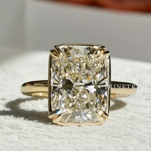 May include: A radiant-cut diamond ring with a gold band. The large, rectangular diamond has a warm, champagne hue and is set in a bezel setting. The ring is displayed on a white surface, with a blurred background.
