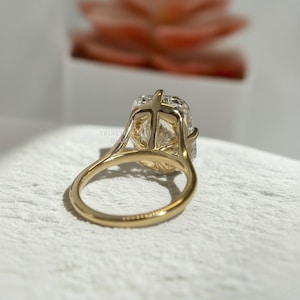 May include: A gold ring featuring a large, rectangular, faceted clear gemstone held in a high-profile, open-work basket setting. The band is smooth and polished, with the text TRINETTE DIAMOND faintly visible on the inner surface of the ring.