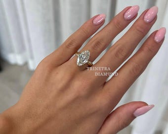 Könnte beinhalten: Ein goldener Ring mit einem großen Diamanten im Marquiseschliff. Der Ring befindet sich an einer Hand mit rosa Nagellack. Der Text "TRINETRA DIAMOND" ist im Bild zu sehen.