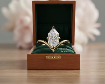 May include: A marquise-cut diamond engagement ring in a wooden box with a green velvet interior. The ring has a unique gold band design. The box is open, revealing the ring. The text "TRINETRA DIAMOND" is visible on the box.