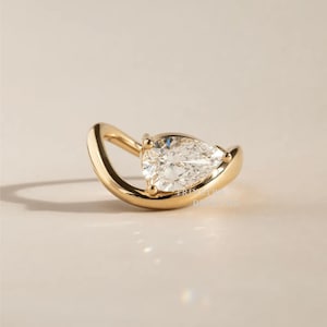 May include: A gold-colored ear cuff featuring a pear-shaped diamond. The diamond is set in a bezel setting, and the cuff curves around the ear. The jewelry is displayed on a neutral background.