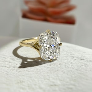 May include: A gold ring featuring a large, rectangular cushion-cut diamond held by four prongs. The band is smooth and polished gold, with a split shank design that connects to the central stone setting. The text TRINITY DIAMOND is visible.