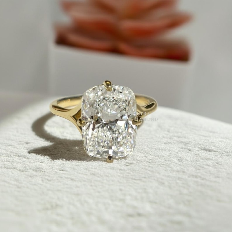 May include: A gold ring featuring a large, rectangular cushion-cut diamond set in a four-prong mounting. The band is smooth and polished, tapering slightly as it meets the central stone. The diamond displays clear facets and high brilliance.