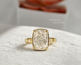 May include: A gold ring featuring a large cushion-cut center stone with a faceted surface, held by four prongs. The band is accented with two smaller round stones on each side. The text TRINETRA DIAMOND is visible in the background.