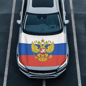 Puede incluir: Un coche plateado con el diseño de la bandera rusa en el capó. La bandera presenta tres franjas horizontales: blanca en la parte superior, azul en el medio y roja en la parte inferior. El emblema de un águila bicéfala está centrado en la franja azul.