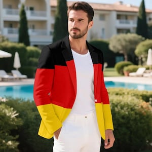 Può includere: Un blazer nero, rosso e giallo, con i colori disposti a bande orizzontali, è indossato sopra una camicia bianca e abbinato a pantaloni bianchi. Il design del blazer rispecchia la bandiera tedesca. Lo sfondo presenta una piscina e una vegetazione lussureggiante.