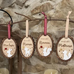Peut inclure: Quatre décorations ovales en bois à bords festonnés, chacune ornée d'une silhouette d'animal blanche, d'une petite étoile ou d'un cœur, et du texte Joyeux Noel avec les noms Mon lapin, Maman, Nounou et Maitresse, suspendues par des rubans.