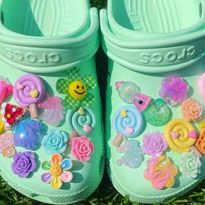 Può includere: Un paio di Crocs verdi decorate con ciondoli colorati, tra cui lecca-lecca, fiori e altri design fantasiosi.