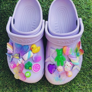 Puede incluir: Un par de zuecos lavanda adornados con abalorios de colores. Los abalorios incluyen mariposas, corazones, un pato amarillo, un oso verde y una figura de Hello Kitty. Los zuecos están sobre hierba verde.
