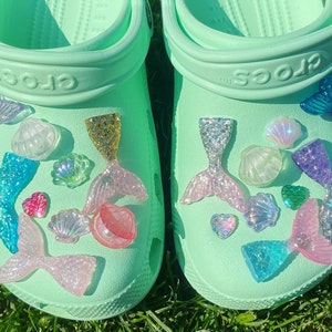 May include: A pair of green Crocs decorated with mermaid tails, seashells, and other ocean-themed charms.