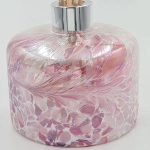 May include: A decorative glass diffuser bottle with a silver collar and wooden reeds. The bottle has a pink and white floral pattern. The reeds are light brown. The diffuser is designed for home fragrance.