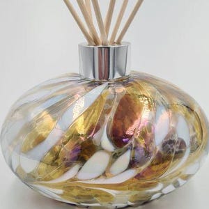 May include: An oval glass diffuser bottle with a silver neck and wooden reeds. The bottle features a swirling pattern of gold, white, and iridescent colours. The reeds are light brown and extend upwards from the bottle.