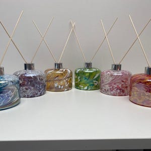 May include: Six decorative glass diffuser bottles in various colours, including blue, purple, gold, green, pink, and red. Each bottle has a silver cap and wooden reeds for fragrance diffusion. The bottles are arranged on a white surface.
