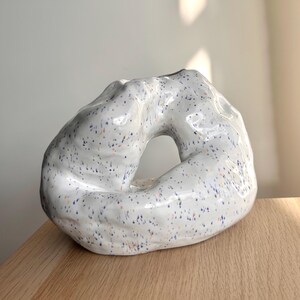 May include: A white ceramic vase with an unusual, organic form. The vase features a large aperture and is speckled with blue, orange, and purple dots. It rests on a wooden surface.