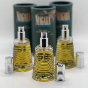 May include: Three glass perfume bottles with silver sprayers and caps, filled with yellow liquid. The bottles are labeled "Migara" and are accompanied by three green cylindrical boxes with the same label. The bottles have a ribbed design.