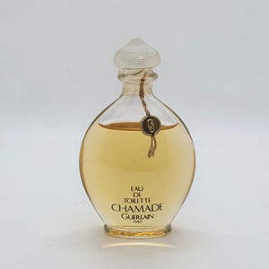 May include: A clear glass perfume bottle with a rounded body containing amber liquid. The bottle features a textured clear stopper and a small gold-toned circular tag tied around the neck with a thin cord. Text reads EAU DE TOILETTE CHAMADE GUERLAIN PARIS.